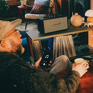 A person sits holding a phone with a music app open, facing a Stanmore IV Black speaker on a shelf filled with vinyl records, in a cozy, retro-styled living room.
