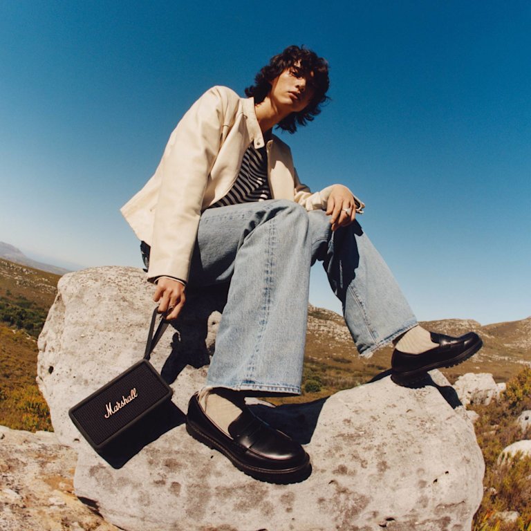 마샬 미들턴 블랙 앤 브라스 휴대용 스피커를 들고 바위 위에 앉아 있는 한 남자.