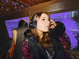 A woman wearing Major V Hendrix Edition headphones sits by a window in a vehicle with a purple-tinted background outside.
