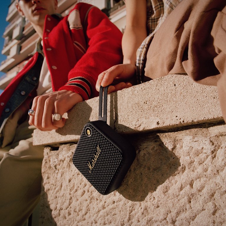 A man and woman sitting on a wall with a Marshall portable speaker called "Willen Black and Brass.