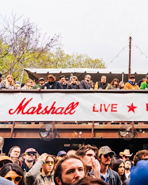 A crowd of people at an outdoor event stands below a balcony with more attendees. A "Marshall Live" banner hangs from the balcony.