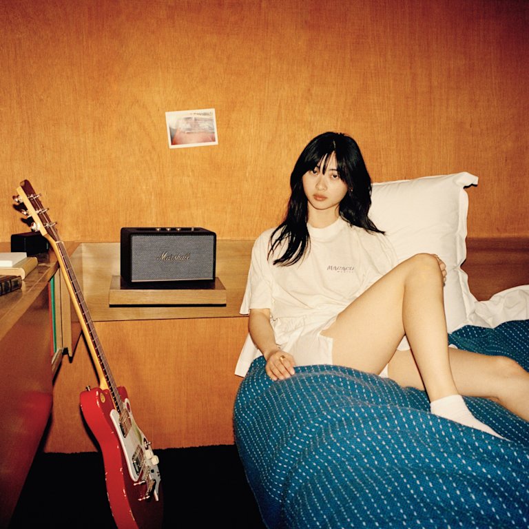 A girl sitting on a bed next to a Marshall ACTON III BLACK guitar.