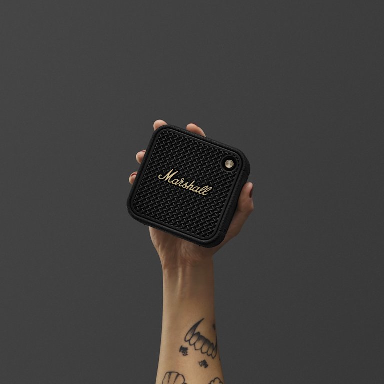 A person holds Marshall Willen II Black and Brass portable bluetooth speaker against a dark background.