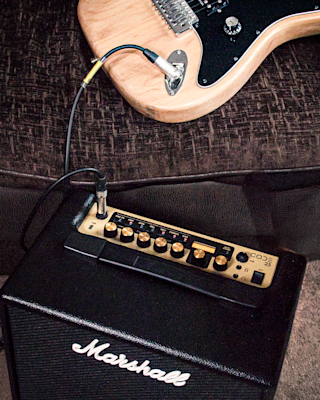 An electric guitar is plugged into a Marshall amplifier, both resting on a carpeted floor next to a brown upholstered couch.