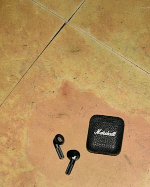 Two black wireless Minor IV earbuds and a black Marshall-branded charging case are placed on a worn, tan tiled floor.