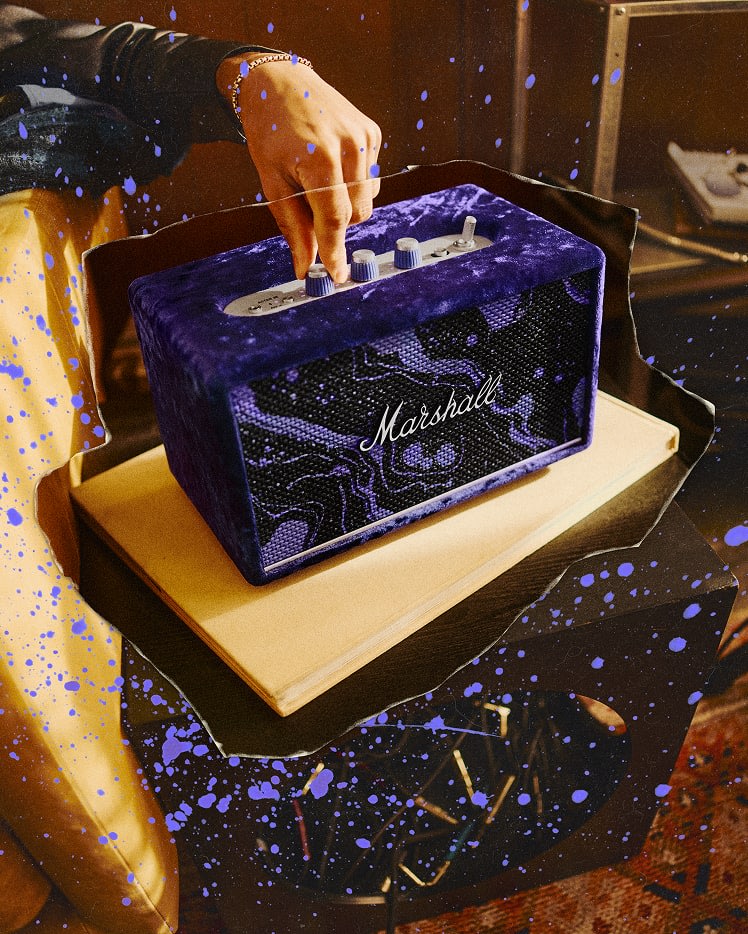 A hand adjusts the knob on a Acton III Marshall x Hendrix 60th Anniversary Edition speaker placed on a book, with blue paint splatters visible across the image.