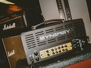 Une tête d'amplificateur de guitare Synergy est placée devant une enceinte d'ampli Marshall, montrant divers boutons de contrôle et prises d'entrée.