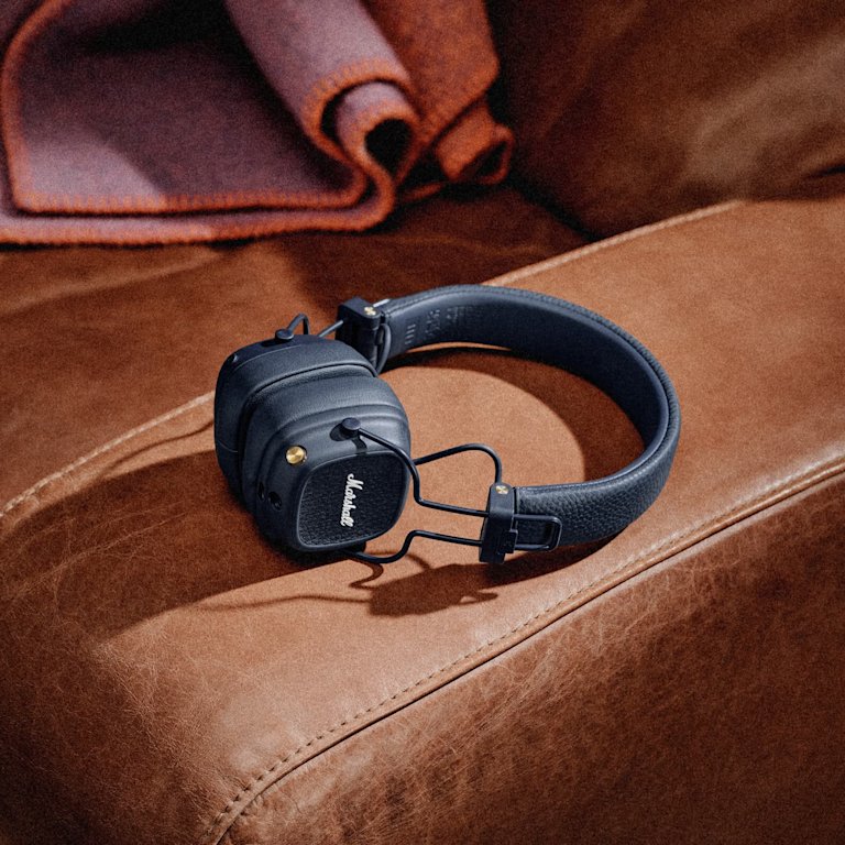 A pair of Marshall Major V Midnight Blue Bluetooth headphones sits on a brown leather couch with a folded blanket in the background.