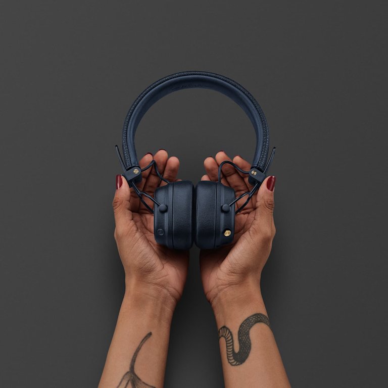 Tattoed hands hold a pair of Marshall Major V Midnight Blue headphones against a plain dark background.