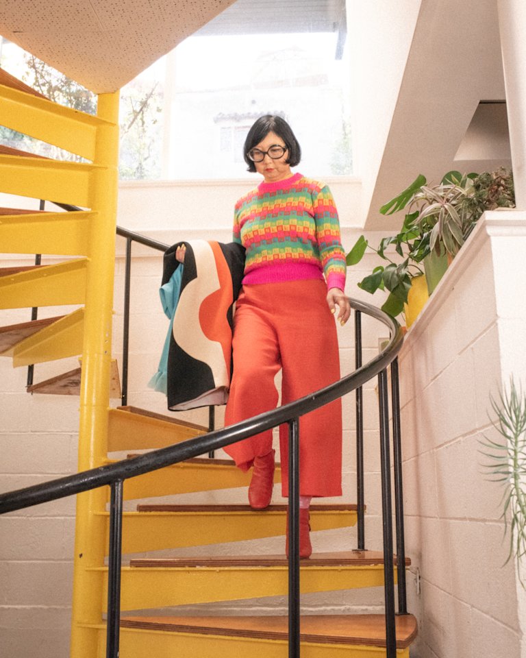 Shirley Kurata walking up a yellow staircase in a colorful sweater.