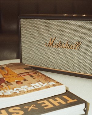 A Marshall Stanmore III Bluetooth speaker sits on a table next to two stacked books, one partially opened, in a softly lit indoor setting.