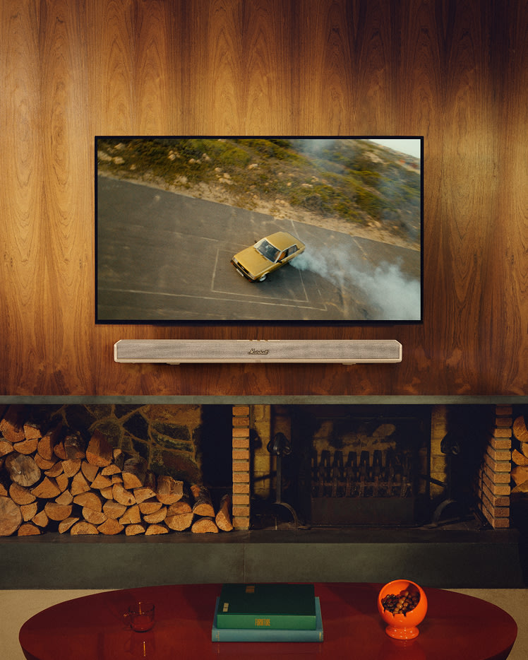 Un televisor colgado en una pared revestida de madera sobre una chimenea, con la barra de sonido Heston 120 Cream debajo de la pantalla y una mesa de centro en primer plano.