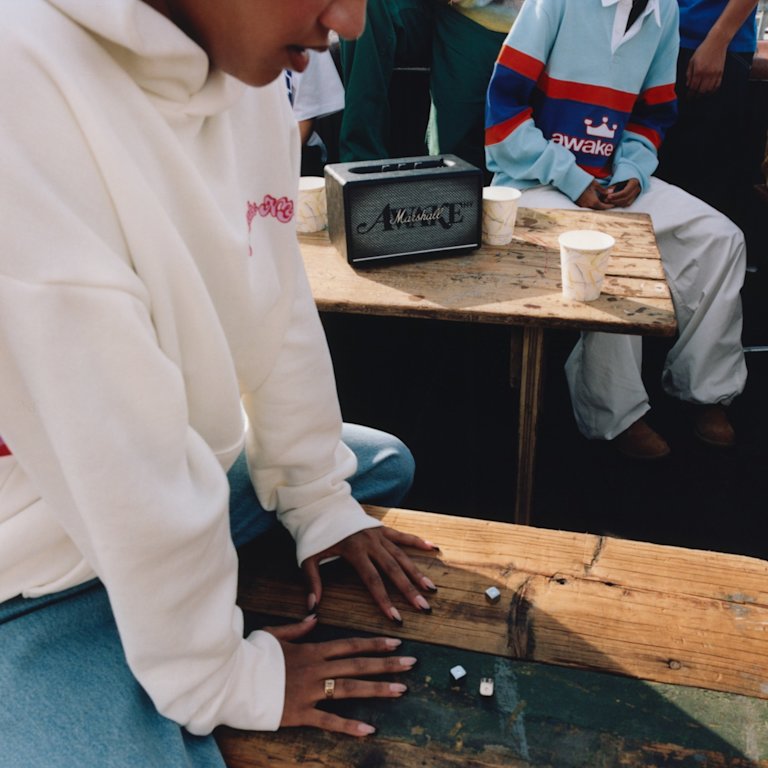 A person in a white hoodie sits on a wooden bench with Acton III Awake NY Edition speaker by Marshall and paper cups in the background.