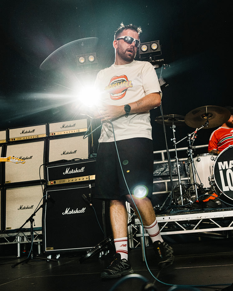 A man wearing sunglasses and a white Mouth Reissue T-shirt performs on stage in front of a stack of Marshall amplifiers and a drum kit.