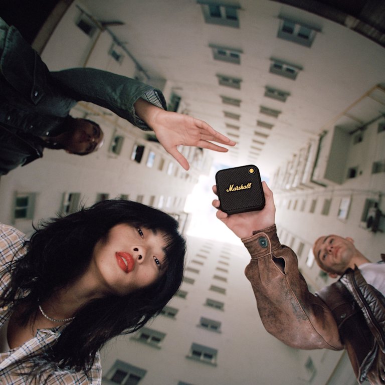 A group of people standing in a building, enjoying the Marshall Willen Black and Brass wireless speaker.