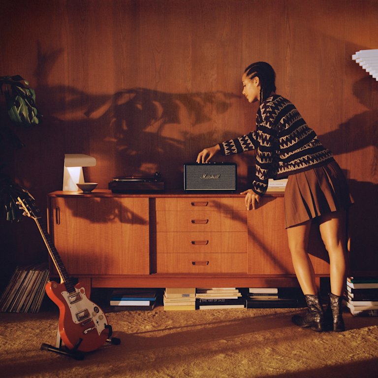 A woman standing next to a black Marshall Stanmore III Black speaker in a room.