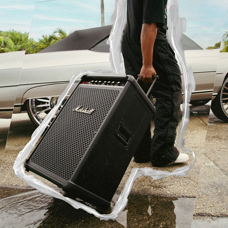 A person pulls a Marshall Bromley 750 party speaker next to a silver car parked outside.