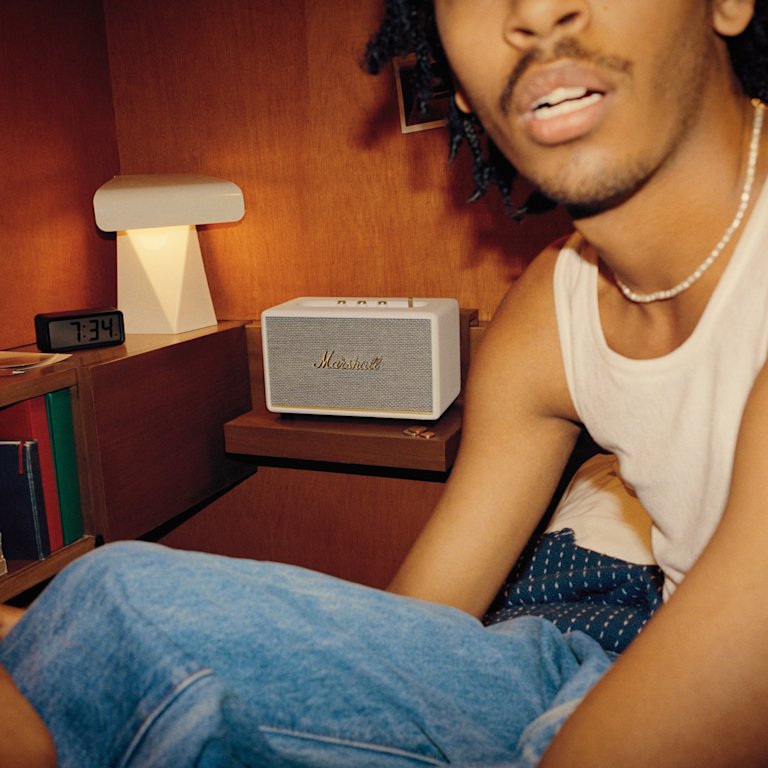 A man with dreadlocks sitting on a bed with a Marshall ACTON III CREAM speaker.