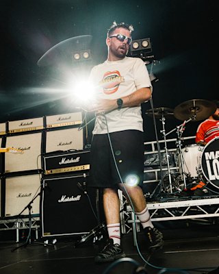 A man wearing Mouth Reissue t-shirt on stage next to a stack of Marshall amplifiers and a drum kit.