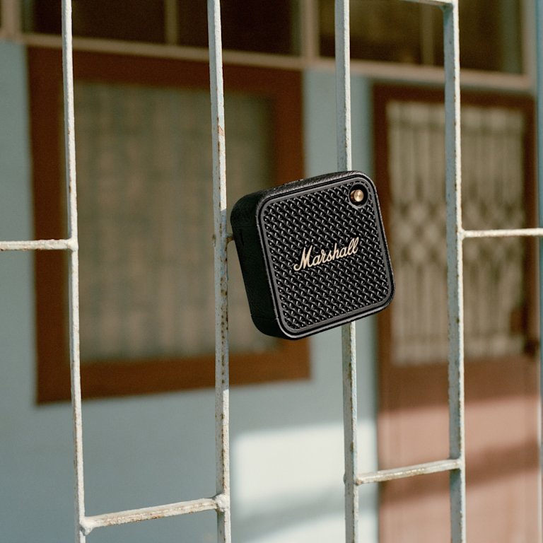 Marshall Willen II Black and Brass portable speaker with the iconic logo is hanging on a metal fence.