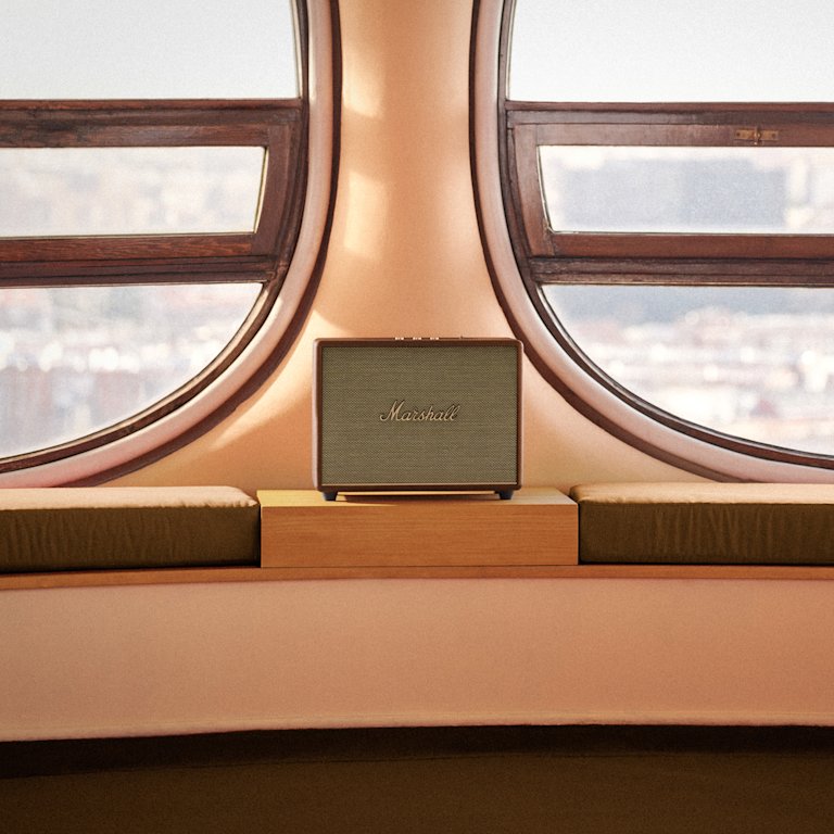 Marshall Woburn III Brown home speaker sits on a wooden shelf between two rounded windows, overlooking a blurred landscape.