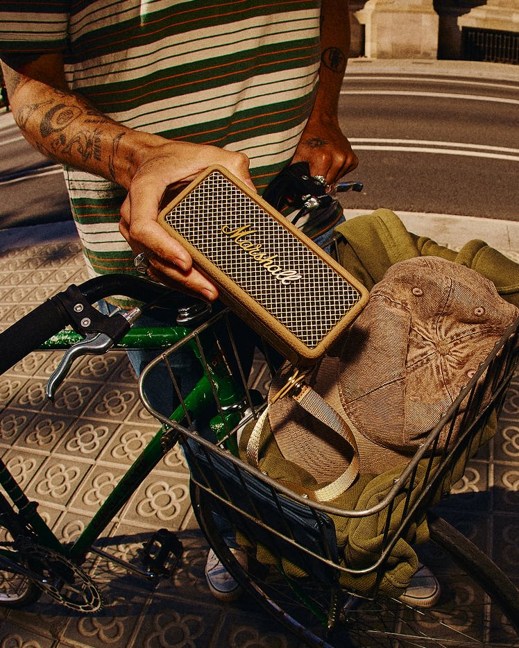 Person holding a Marshall Emberton III Brown portable speaker while standing next to a green bicycle with a khaki bag and brown cap in the front basket.