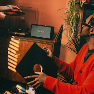 Two people handling vinyl records near a wooden cabinet, with Acton IV Black speaker speaker in the background.
