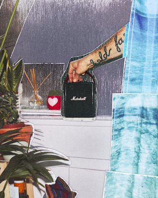 A person adjusting Marshall Stockwell III Black and Brass speaker placed next to a red heart candle, plants, and blue fabric patterns in the background.
