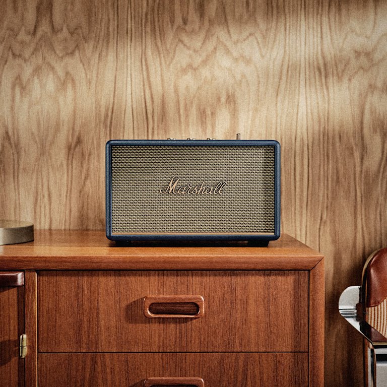 Marshall Acton III Midnight Blue Bluetooth speaker sits on a wooden cabinet against a wood-paneled wall.