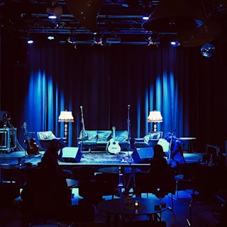 A dimly lit stage with sofas, a rug, floor lamps, and guitars under blue lighting; audience members are seated at tables in front of the stage.
