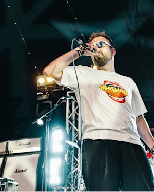 A man wearing a Mouth Reissue t-shirt sings on stage in front of Marshall amplifiers.