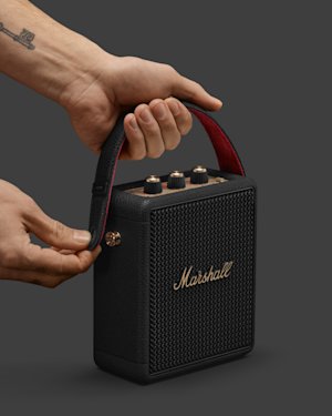 Two hands hold a portable Marshall speaker by its leather strap, adjusting the metal button on the side. The speaker is black with gold accents and control knobs on top.