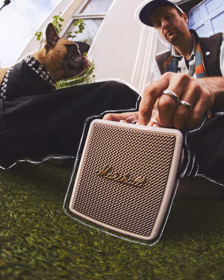 A man sits on grass next to a dog, adjusting a portable Stockwell III Cream speaker, with a building and greenery in the background.