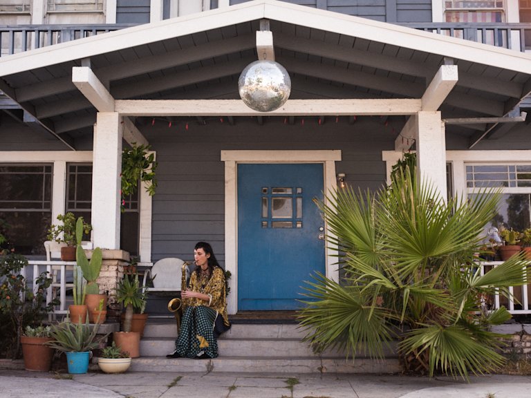 Zumi Rosow sitting on the steps of a house playing a saxophone.