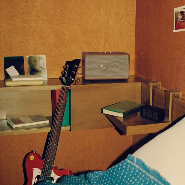 A brown speaker, known as the Marshall Acton III Brown, sits on a bed alongside a guitar.