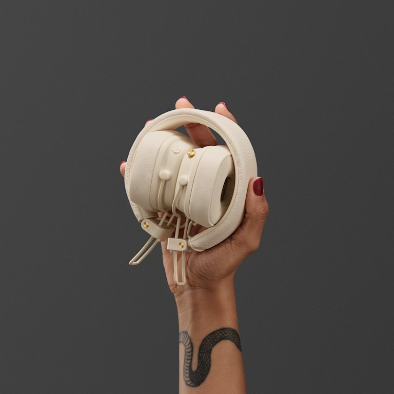 A person with red nail polish and a tattoo on their wrist holds a pair of Marshall Major V Cream on-ear headphones against a gray background.