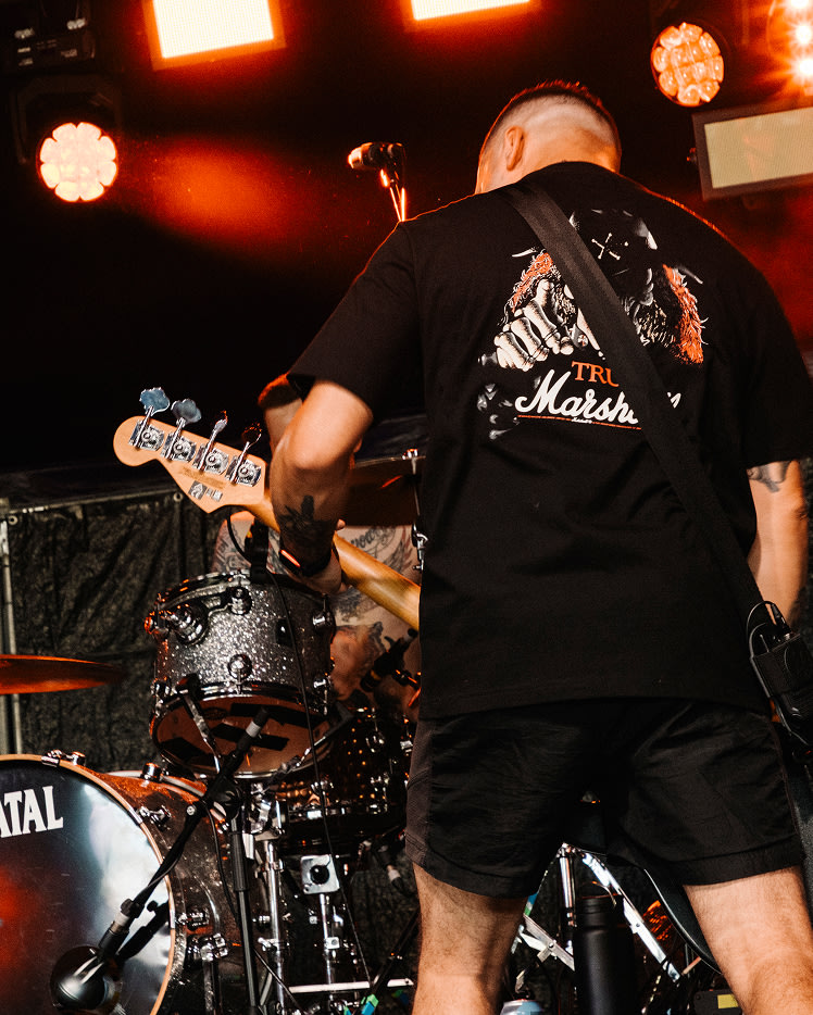 A bassist performing on stage in front of a drum kit, wearing Marshall Stax Reissue T-shirt Black.