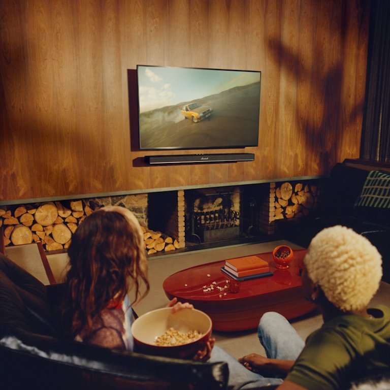 Two people sit on a couch and watching a TV with the Marshall Heston 120 soundbar, in a cozy wood-paneled living room.