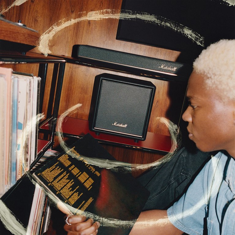 A person with short blonde hair examines a vinyl record sleeve near shelves of records, and a Marshall Heston Sub 200 Black subwoofer.