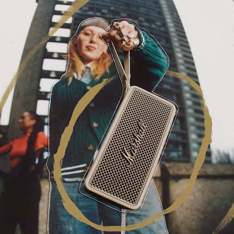 A person holds a Marshall Middleton II Cream portable Bluetooth speaker toward the camera, with a tall building in the background.