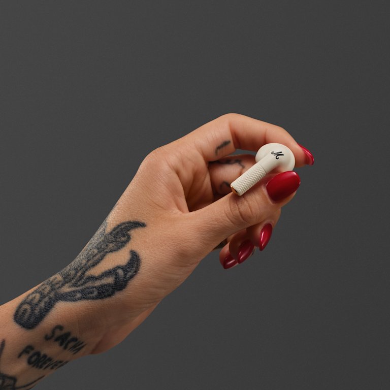 A hand with tattoos and red nails holds a Marshall Minor IV Cream earbud against a dark gray background.