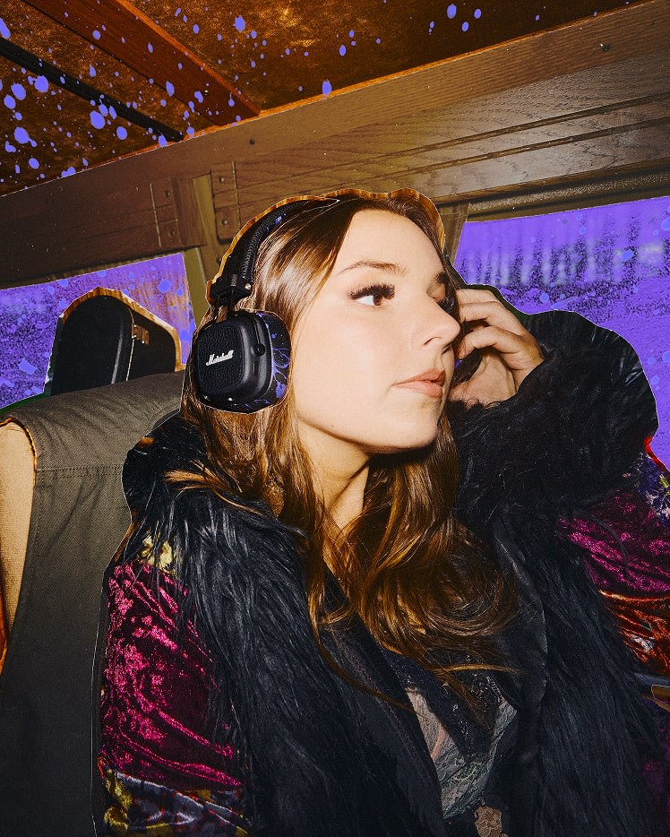 A woman wearing Major V Hendrix Edition headphones sits on a bus with purple lighting and raindrops on the window behind her.
