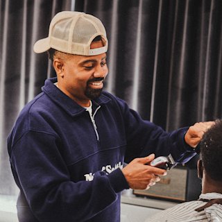 Vince Jamael wearing a cap and navy sweatshirt is smiling while using clippers to cut a client's hair in a barbershop.
