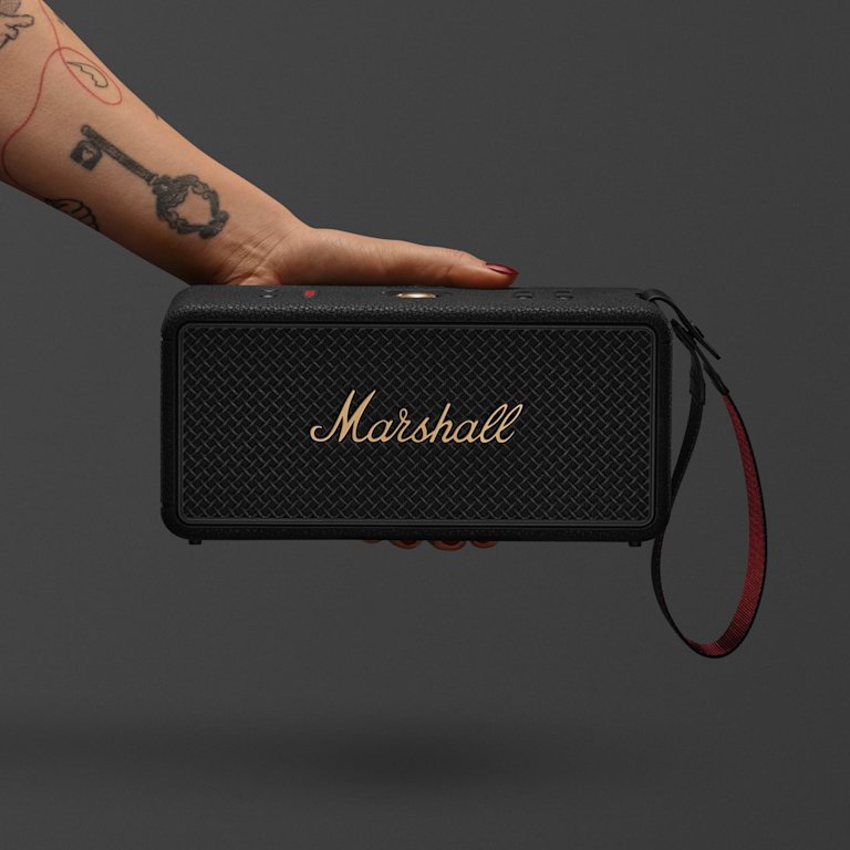 A tattooed hand holds a Marshall Middleton II Black and Brass portable Bluetooth speaker, set against a plain dark background.
