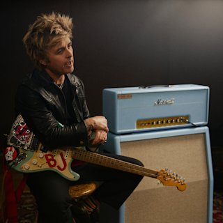 Una foto de Billie Joe Armstrong sentado en un taburete, sosteniendo una guitarra eléctrica con pegatinas, junto al amplificador azul Marshall 1959BJA.