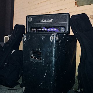 JMH Studio Head sits on a worn black speaker cabinet between two soft guitar cases, against a brick wall background.