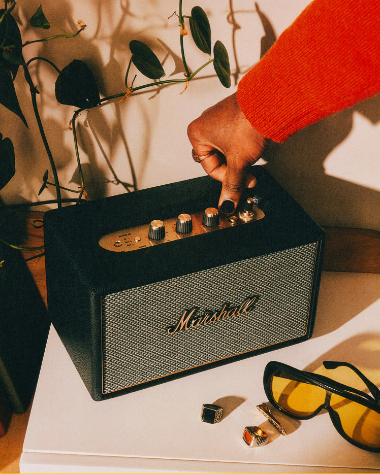 A person in a red sleeve adjusts the volume knob on Marshall Acton IV Black speaker placed on a white surface, near sunglasses and rings.