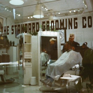 A client sits in a barber chair with a white cape while a barber stands nearby in a well-lit, modern grooming salon, the Standard Grooming Co.