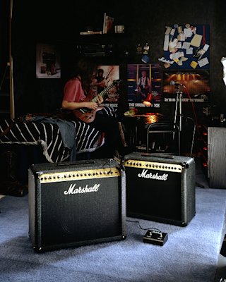 Guitar amplifiers on the floor next to a bed where a person is playing the guitar