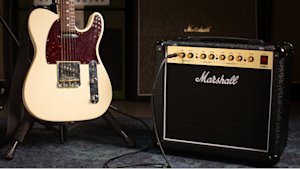 A cream-colored electric guitar on a stand next to a black Marshall amplifier, both placed on a carpeted floor.